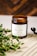 A brown glass jar candle with a white label named 'Verso L'Alto' is placed on a textured beige surface. Green leafy branches are positioned beside the jar, adding a natural element. The background is a warm, out-of-focus wooden texture.