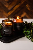Two amber glass candles are set on a dark tray with greenery surrounding them. One candle is lit, casting a warm, cozy glow. The candle labels read 'CHRISMa'. The background features a wooden texture, enhancing the rustic and calming atmosphere.