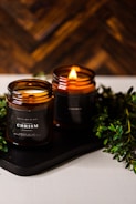 Two amber glass candles are set on a dark tray with greenery surrounding them. One candle is lit, casting a warm, cozy glow. The candle labels read 'CHRISMa'. The background features a wooden texture, enhancing the rustic and calming atmosphere.