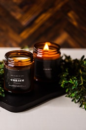 Two amber glass candles are set on a dark tray with greenery surrounding them. One candle is lit, casting a warm, cozy glow. The candle labels read 'CHRISMa'. The background features a wooden texture, enhancing the rustic and calming atmosphere.