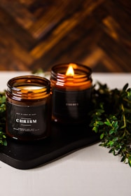 Two amber glass candles are set on a dark tray with greenery surrounding them. One candle is lit, casting a warm, cozy glow. The candle labels read 'CHRISMa'. The background features a wooden texture, enhancing the rustic and calming atmosphere.