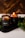 Two amber glass candles are set on a dark tray with greenery surrounding them. One candle is lit, casting a warm, cozy glow. The candle labels read 'CHRISMa'. The background features a wooden texture, enhancing the rustic and calming atmosphere.