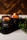 Two amber glass candles are set on a dark tray with greenery surrounding them. One candle is lit, casting a warm, cozy glow. The candle labels read 'CHRISMa'. The background features a wooden texture, enhancing the rustic and calming atmosphere.