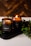 Two amber glass candles are set on a dark tray with greenery surrounding them. One candle is lit, casting a warm, cozy glow. The candle labels read 'CHRISMa'. The background features a wooden texture, enhancing the rustic and calming atmosphere.