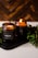 Two amber glass candles are set on a dark tray with greenery surrounding them. One candle is lit, casting a warm, cozy glow. The candle labels read 'CHRISMa'. The background features a wooden texture, enhancing the rustic and calming atmosphere.