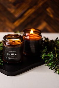 Two amber glass candles are set on a dark tray with greenery surrounding them. One candle is lit, casting a warm, cozy glow. The candle labels read 'CHRISMa'. The background features a wooden texture, enhancing the rustic and calming atmosphere.