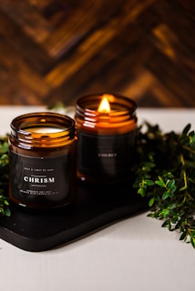 Two amber glass candles are set on a dark tray with greenery surrounding them. One candle is lit, casting a warm, cozy glow. The candle labels read 'CHRISMa'. The background features a wooden texture, enhancing the rustic and calming atmosphere.