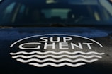 The image features the logo 'SUP Ghent' prominently displayed on a surface, likely the hood of a vehicle. The logo includes stylized waves beneath the text.