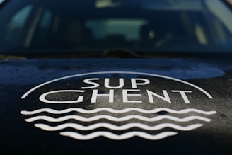 The image features the logo 'SUP Ghent' prominently displayed on a surface, likely the hood of a vehicle. The logo includes stylized waves beneath the text.