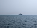 A naval ship is seen on the horizon over a calm ocean, under a clear, expansive sky.