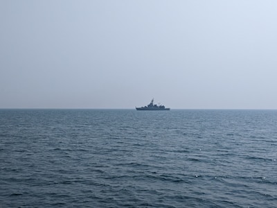 A naval ship is seen on the horizon over a calm ocean, under a clear, expansive sky.