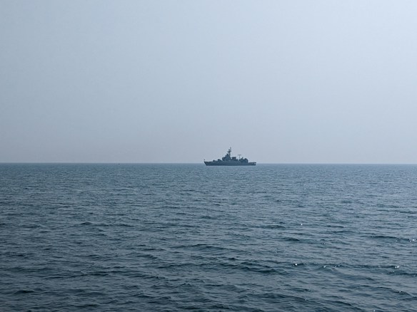 A naval ship is seen on the horizon over a calm ocean, under a clear, expansive sky.