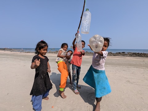 A group of five children are playing on a sandy beach near the ocean. They are holding up empty plastic bottles attached to a stick. The scene is lively and joyful, with clear blue skies and a calm sea in the background. The children are dressed colorfully and appear to be having fun.