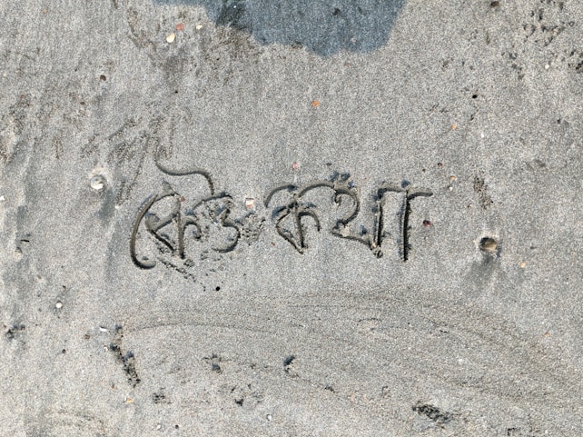 The image displays text written in a script on a sandy surface, with small shells and pebbles scattered around. The sand has a light gray color, and there are some darker patches where moisture might be present.