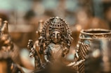 An intricately detailed bronze sculpture of a deity figure highlighted against a blurred background. The artwork features precise engravings and elaborate patterns, capturing the cultural and artistic heritage. The soft lighting accentuates the metallic textures and gives a warm glow.