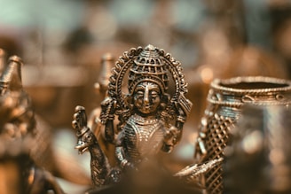 An intricately detailed bronze sculpture of a deity figure highlighted against a blurred background. The artwork features precise engravings and elaborate patterns, capturing the cultural and artistic heritage. The soft lighting accentuates the metallic textures and gives a warm glow.