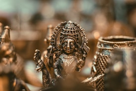 An intricately detailed bronze sculpture of a deity figure highlighted against a blurred background. The artwork features precise engravings and elaborate patterns, capturing the cultural and artistic heritage. The soft lighting accentuates the metallic textures and gives a warm glow.