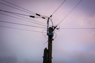 A utility pole stands tall, with multiple power lines extending outward in different directions. The lines are affixed to the pole using various insulators and connectors. The background features a pastel-colored sky that transitions between shades of pink, purple, and blue, suggesting either sunrise or sunset.