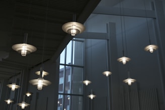 Modern pendant lights with a layered, circular design hang from a high ceiling. The lights are evenly spaced and emit a soft, white glow. Large windows in the backdrop allow natural light to enter, providing a view of a building outside.