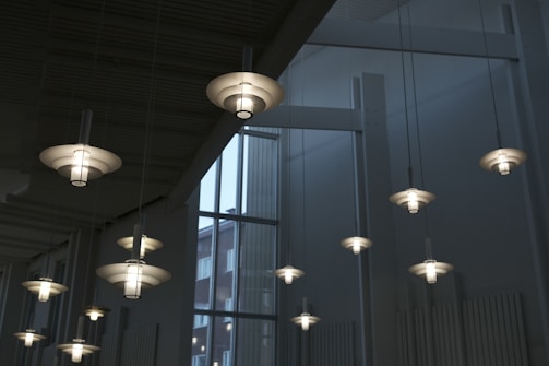 Modern pendant lights with a layered, circular design hang from a high ceiling. The lights are evenly spaced and emit a soft, white glow. Large windows in the backdrop allow natural light to enter, providing a view of a building outside.