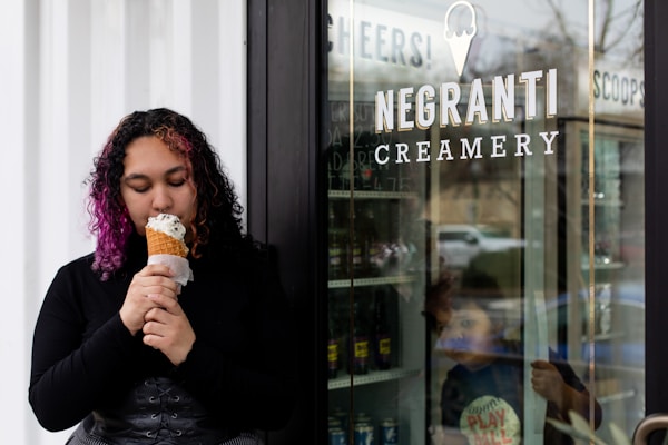 A person with curly hair enjoys a waffle cone with ice cream outside a store, with 'Negranti Creamery' sign on the glass door. A child looks out from inside the store, which has reflective glass showing part of the street outside.
