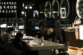 A dimly lit café interior features multiple people sitting at tables with laptops and phones. Large circular mirrors or artwork are on the wall, and a counter with pastries is in the foreground. Ambient lighting creates a cozy atmosphere, with reflections in the glass wall showing part of the café logo.