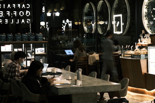 A dimly lit café interior features multiple people sitting at tables with laptops and phones. Large circular mirrors or artwork are on the wall, and a counter with pastries is in the foreground. Ambient lighting creates a cozy atmosphere, with reflections in the glass wall showing part of the café logo.