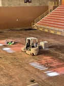A construction scene inside a large indoor space features a forklift parked on the dusty floor. Near it, there are several bags of materials and a stack of bricks. An array of stadium-style seating with red chairs is visible, leading up flights of stairs. The lighting casts sporadic bright patches on the floor.