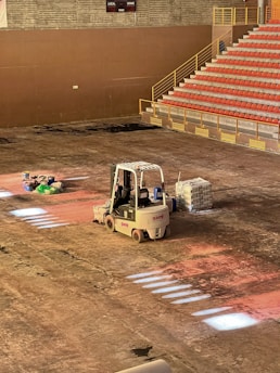 A construction scene inside a large indoor space features a forklift parked on the dusty floor. Near it, there are several bags of materials and a stack of bricks. An array of stadium-style seating with red chairs is visible, leading up flights of stairs. The lighting casts sporadic bright patches on the floor.