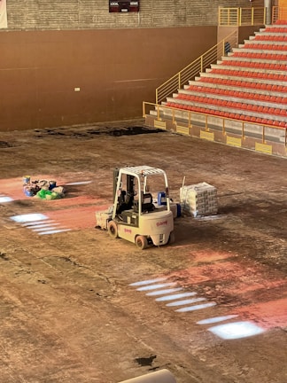 A construction scene inside a large indoor space features a forklift parked on the dusty floor. Near it, there are several bags of materials and a stack of bricks. An array of stadium-style seating with red chairs is visible, leading up flights of stairs. The lighting casts sporadic bright patches on the floor.