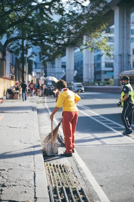 A person wearing a yellow shirt and red pants is sweeping the street with a broom. The scene is set on a sunny day under a tree, with an elevated train track in the background. Several pedestrians are walking along the sidewalk, and a person in a safety uniform is nearby.