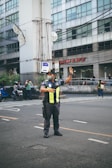 A traffic officer stands on the road directing vehicles with one arm raised. He is wearing a uniform with reflective yellow stripes and a face mask. In the background, there is a large building with multiple windows and people on motorcycles passing by.
