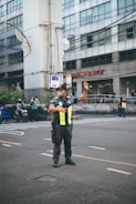 A traffic officer stands on the road directing vehicles with one arm raised. He is wearing a uniform with reflective yellow stripes and a face mask. In the background, there is a large building with multiple windows and people on motorcycles passing by.