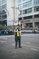 A traffic officer stands on the road directing vehicles with one arm raised. He is wearing a uniform with reflective yellow stripes and a face mask. In the background, there is a large building with multiple windows and people on motorcycles passing by.