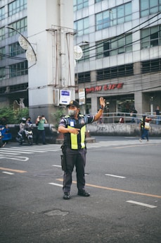 A traffic officer stands on the road directing vehicles with one arm raised. He is wearing a uniform with reflective yellow stripes and a face mask. In the background, there is a large building with multiple windows and people on motorcycles passing by.