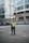 A traffic officer stands on the road directing vehicles with one arm raised. He is wearing a uniform with reflective yellow stripes and a face mask. In the background, there is a large building with multiple windows and people on motorcycles passing by.