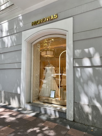 A store window display featuring a white bridal gown on a mannequin. The store's exterior is painted light gray with the brand name 'PRONOVIAS' prominently displayed above the window in gold letters. Shadows from nearby trees create a pattern on the sidewalk and building wall.