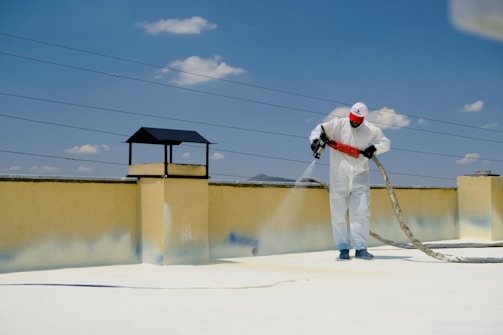 A person wearing protective gear is applying a white substance on a flat surface, possibly for insulation or coating. The individual is on a rooftop with a clear blue sky overhead. There are power lines in the background and a structure resembling a chimney.