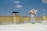 A person wearing protective gear is applying a white substance on a flat surface, possibly for insulation or coating. The individual is on a rooftop with a clear blue sky overhead. There are power lines in the background and a structure resembling a chimney.