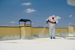 A person wearing protective gear is applying a white substance on a flat surface, possibly for insulation or coating. The individual is on a rooftop with a clear blue sky overhead. There are power lines in the background and a structure resembling a chimney.