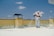 A person wearing protective gear is applying a white substance on a flat surface, possibly for insulation or coating. The individual is on a rooftop with a clear blue sky overhead. There are power lines in the background and a structure resembling a chimney.