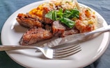 A plate of food featuring grilled meat pieces, accompanied by a creamy vegetable salad with visible carrots and peas. There is also a serving of mashed sweet potatoes on the side, topped with fresh green herbs. A fork and knife are placed on the edge of the white plate, which rests on a dark-colored tablecloth.