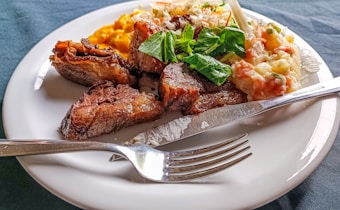 A plate of food featuring grilled meat pieces, accompanied by a creamy vegetable salad with visible carrots and peas. There is also a serving of mashed sweet potatoes on the side, topped with fresh green herbs. A fork and knife are placed on the edge of the white plate, which rests on a dark-colored tablecloth.