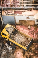 A yellow truck filled with rubble is parked in a yard surrounded by debris. The ground is covered in dirt and scattered pieces of construction material. Two people are interacting with the debris, engaged in some form of manual labor. The scene is rustic and chaotic, with worn-out surfaces visible on the surrounding walls.