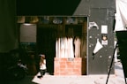 A small child in a helmet stands in front of a clothing shop. The shop features hanging white garments, and its entrance is framed with brick. The child's shadow is cast on the pavement, and the shop is partly in shadow with sunlight streaming in.