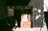 A small child in a helmet stands in front of a clothing shop. The shop features hanging white garments, and its entrance is framed with brick. The child's shadow is cast on the pavement, and the shop is partly in shadow with sunlight streaming in.