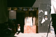 A small child in a helmet stands in front of a clothing shop. The shop features hanging white garments, and its entrance is framed with brick. The child's shadow is cast on the pavement, and the shop is partly in shadow with sunlight streaming in.