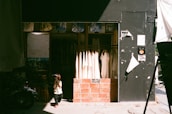 A small child in a helmet stands in front of a clothing shop. The shop features hanging white garments, and its entrance is framed with brick. The child's shadow is cast on the pavement, and the shop is partly in shadow with sunlight streaming in.