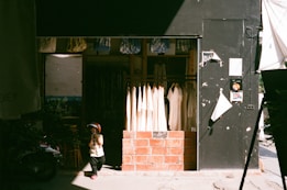 A small child in a helmet stands in front of a clothing shop. The shop features hanging white garments, and its entrance is framed with brick. The child's shadow is cast on the pavement, and the shop is partly in shadow with sunlight streaming in.