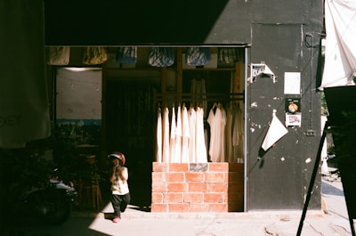 A small child in a helmet stands in front of a clothing shop. The shop features hanging white garments, and its entrance is framed with brick. The child's shadow is cast on the pavement, and the shop is partly in shadow with sunlight streaming in.